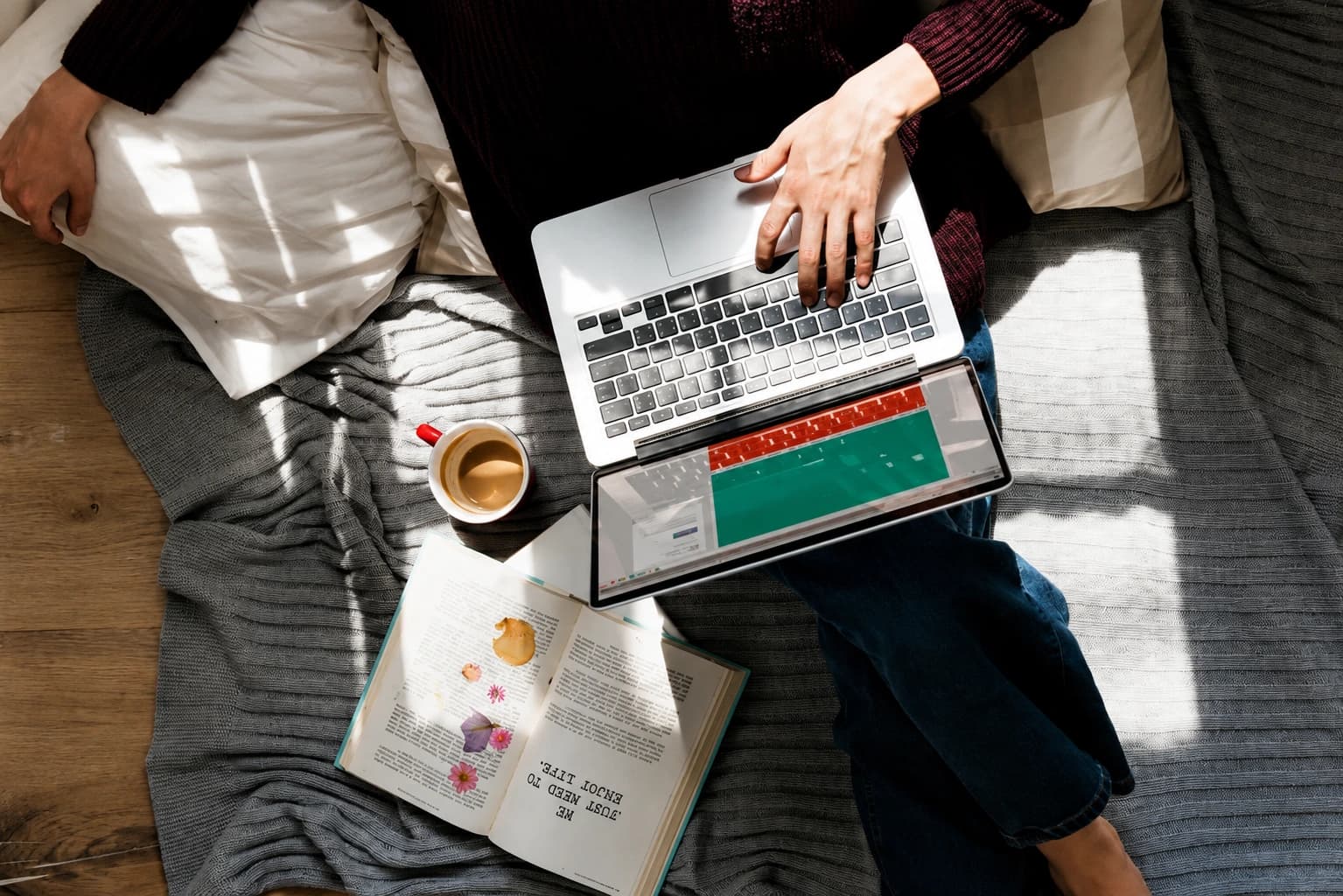 Top-down view of a person on a gray sofa using a laptop, with a tablet on the lap, an open book nearby, and a coffee mug nearby.