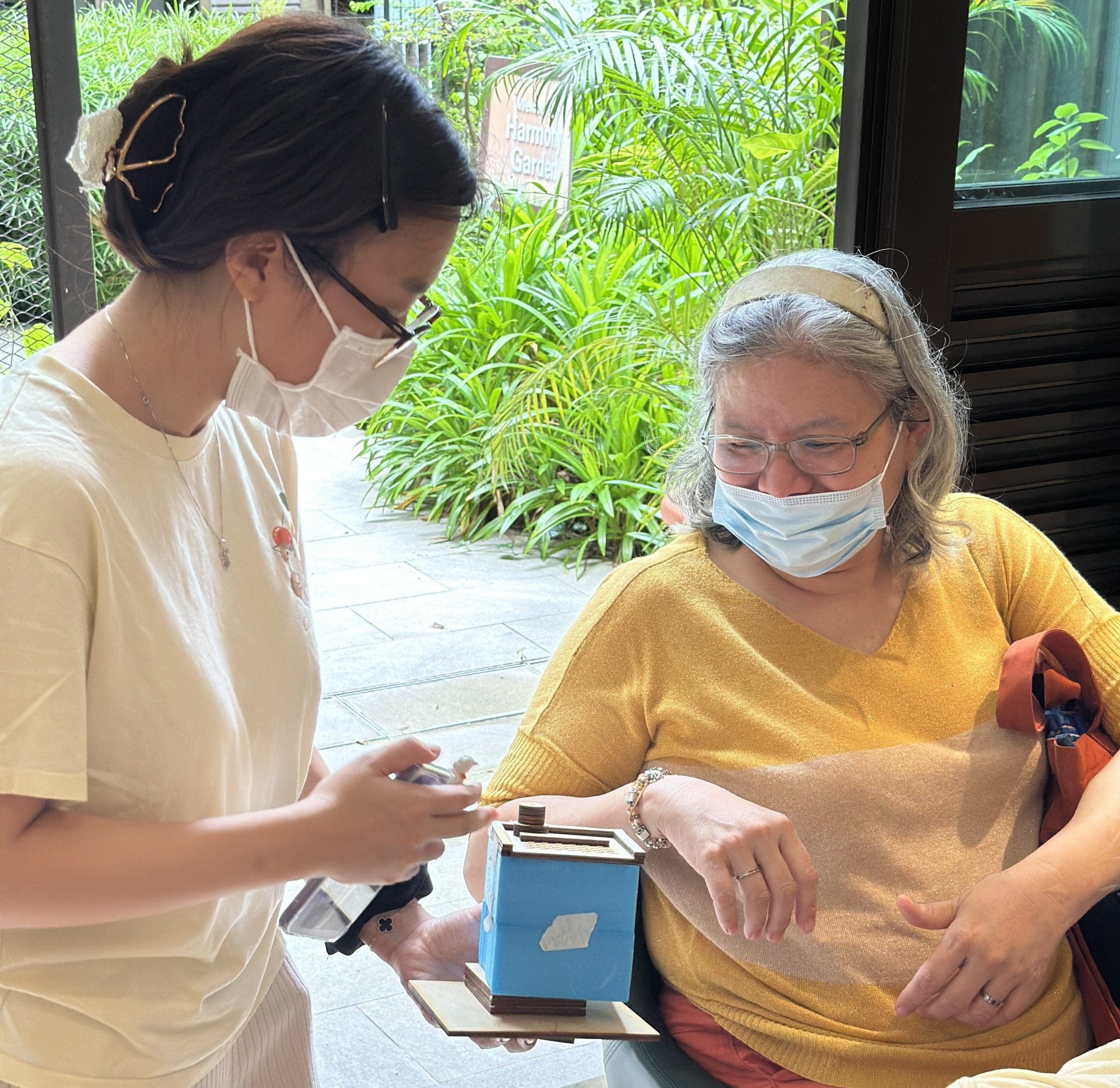 Two women wearing face masks interacting indoors near a window overlooking a garden, with one woman holding a small blue-and-wood model structure on a base.