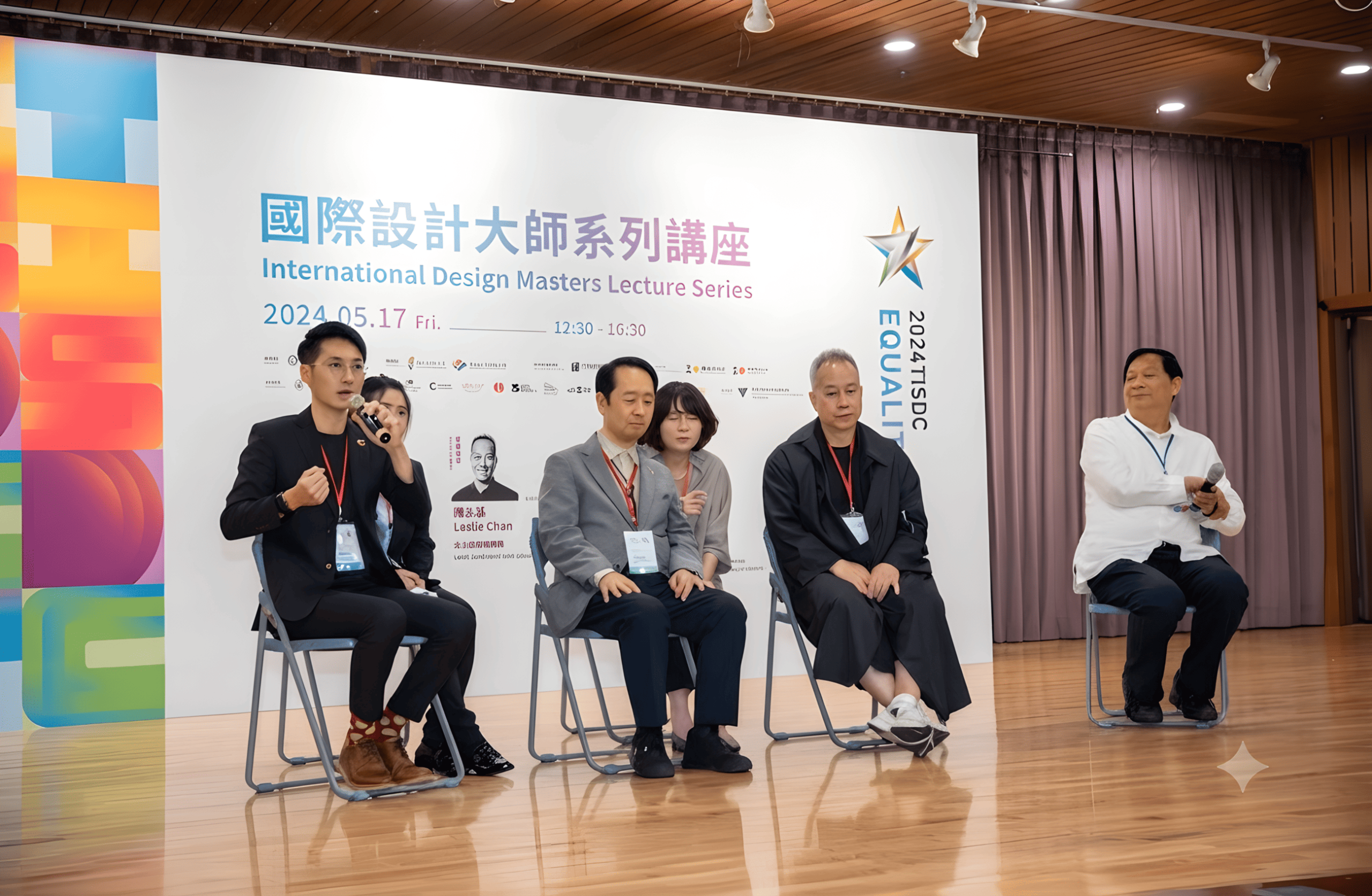 Five panelists seated on a stage during a lecture, with a banner reading 'International Design Masters Lecture Series' behind them and a presenter holding a microphone on the left side.