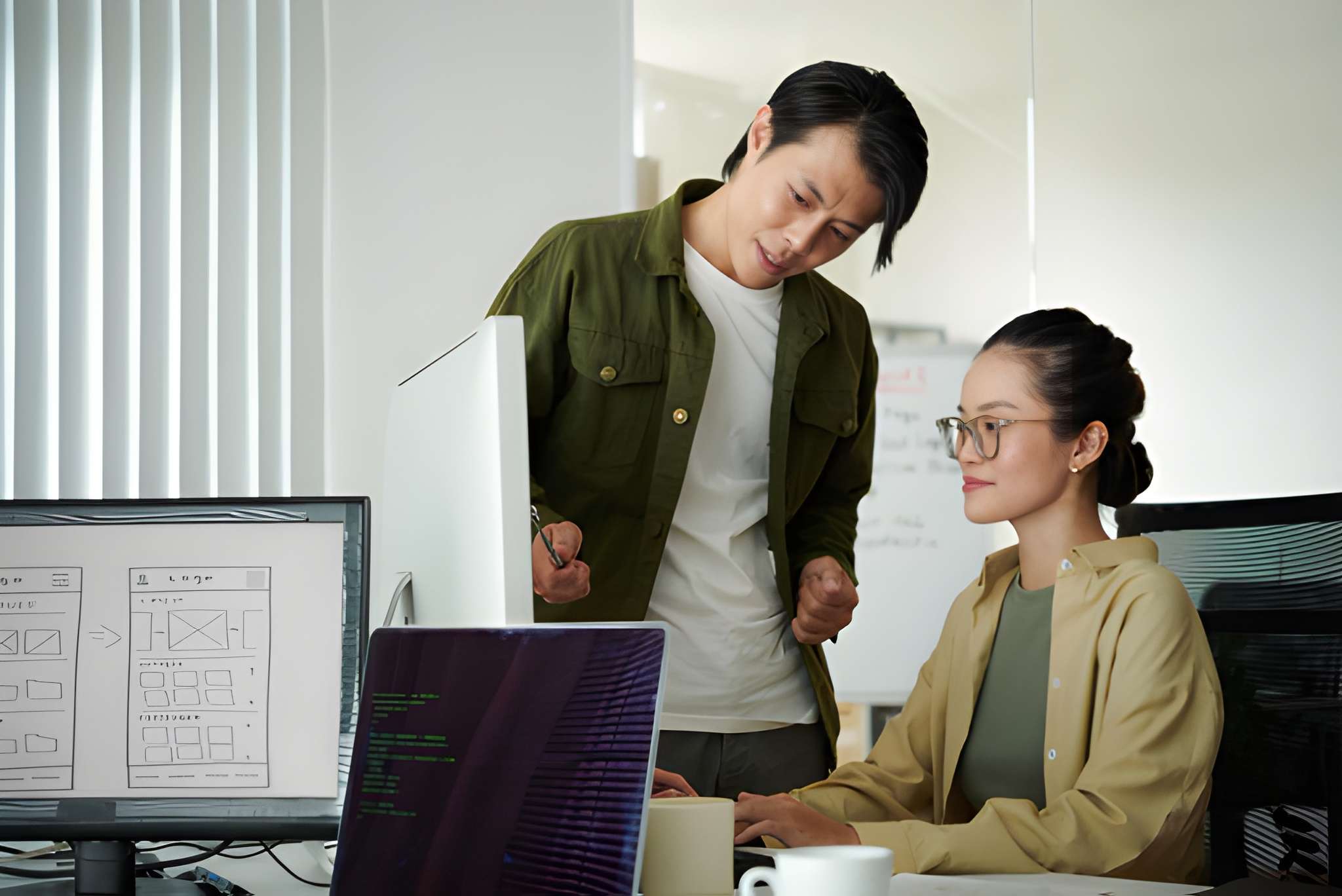 Two colleagues collaborate at a desk: a standing woman points at a monitor while a seated coworker looks on.