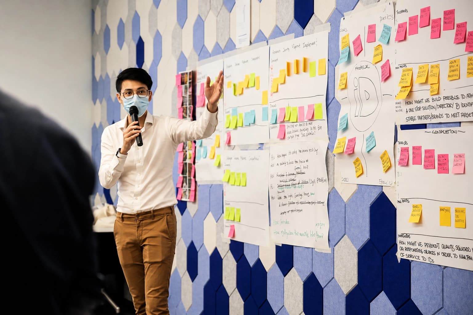 Presenter with a mask speaks into a microphone while explaining colorful sticky-note charts on a wall workshop setup.