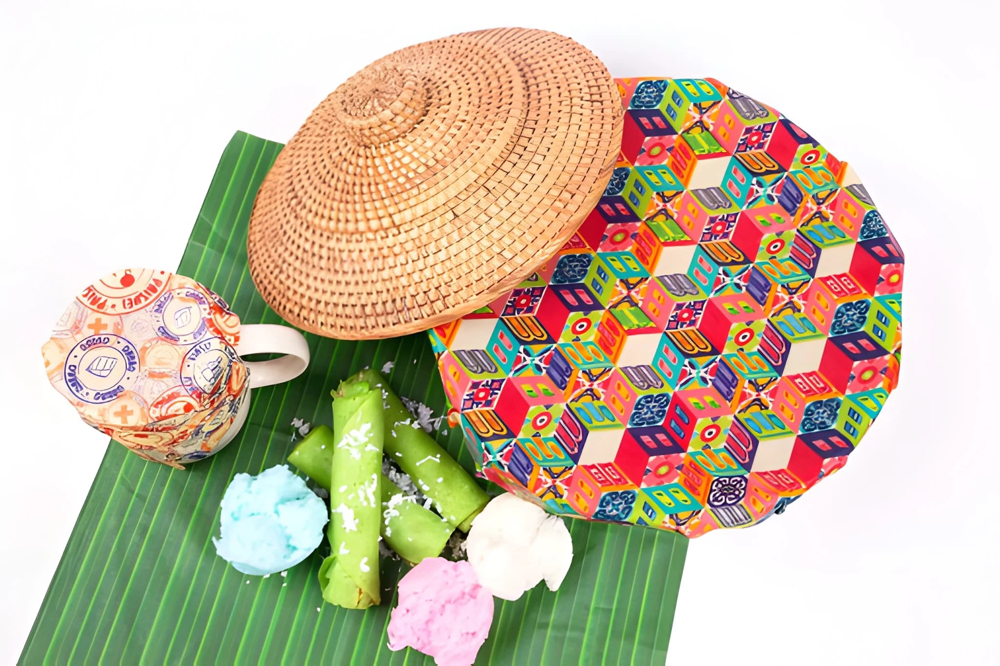 Colorful Asian snacks, green rolled crepes, and pastel cakes arranged on a banana leaf with woven containers and a patterned mug.
