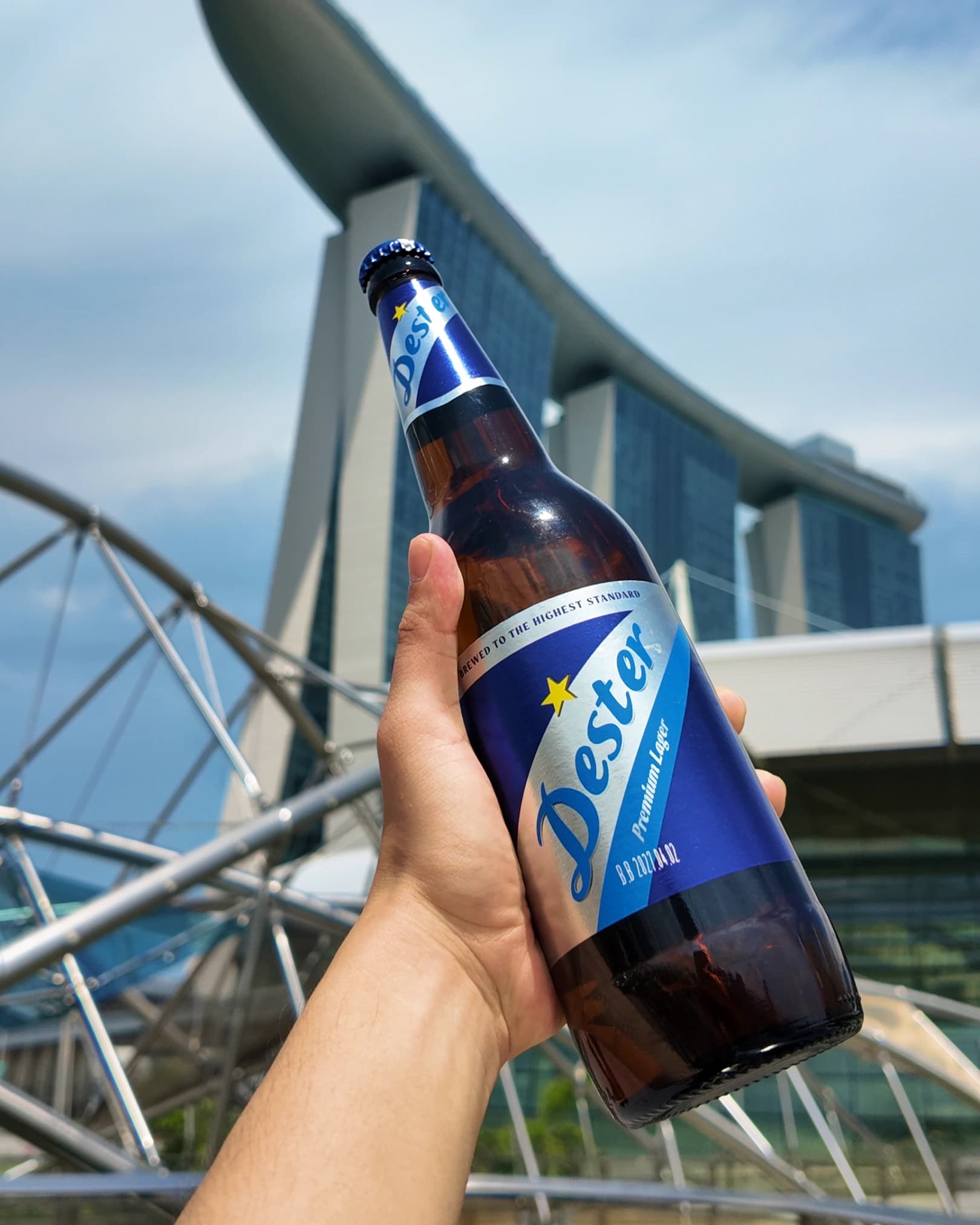 Hand holding a Desperados beer bottle with a modern steel-and-glass bridge in the background.