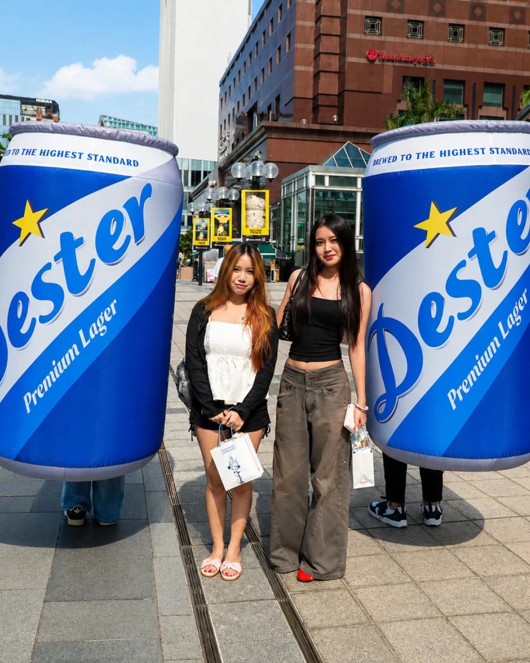 Two young women pose between oversized inflatable beer cans labeled 'Premium Lager' in a city street.