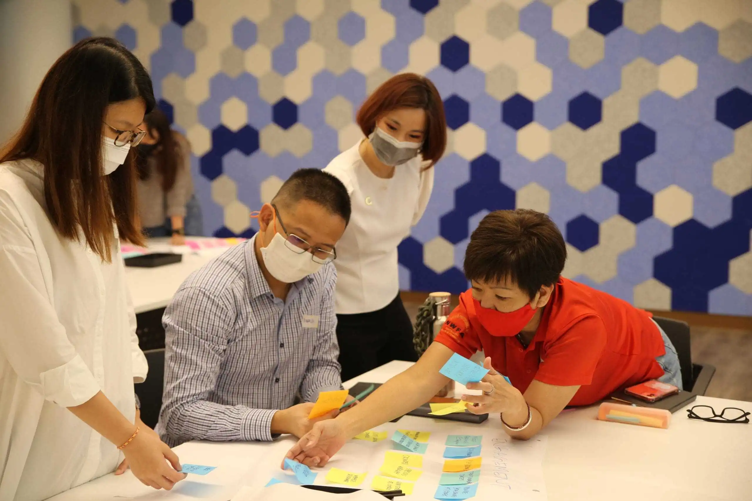 Group of people wearing masks around a table, placing sticky notes on a large sheet of paper.